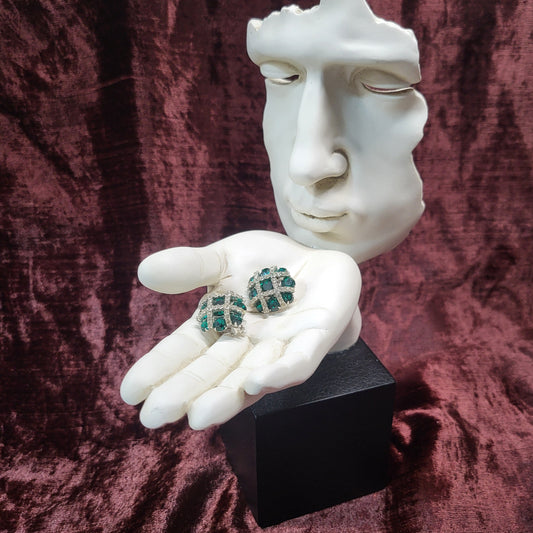 White mannequin hands holding a black box with green gemstone earrings on a maroon fabric background